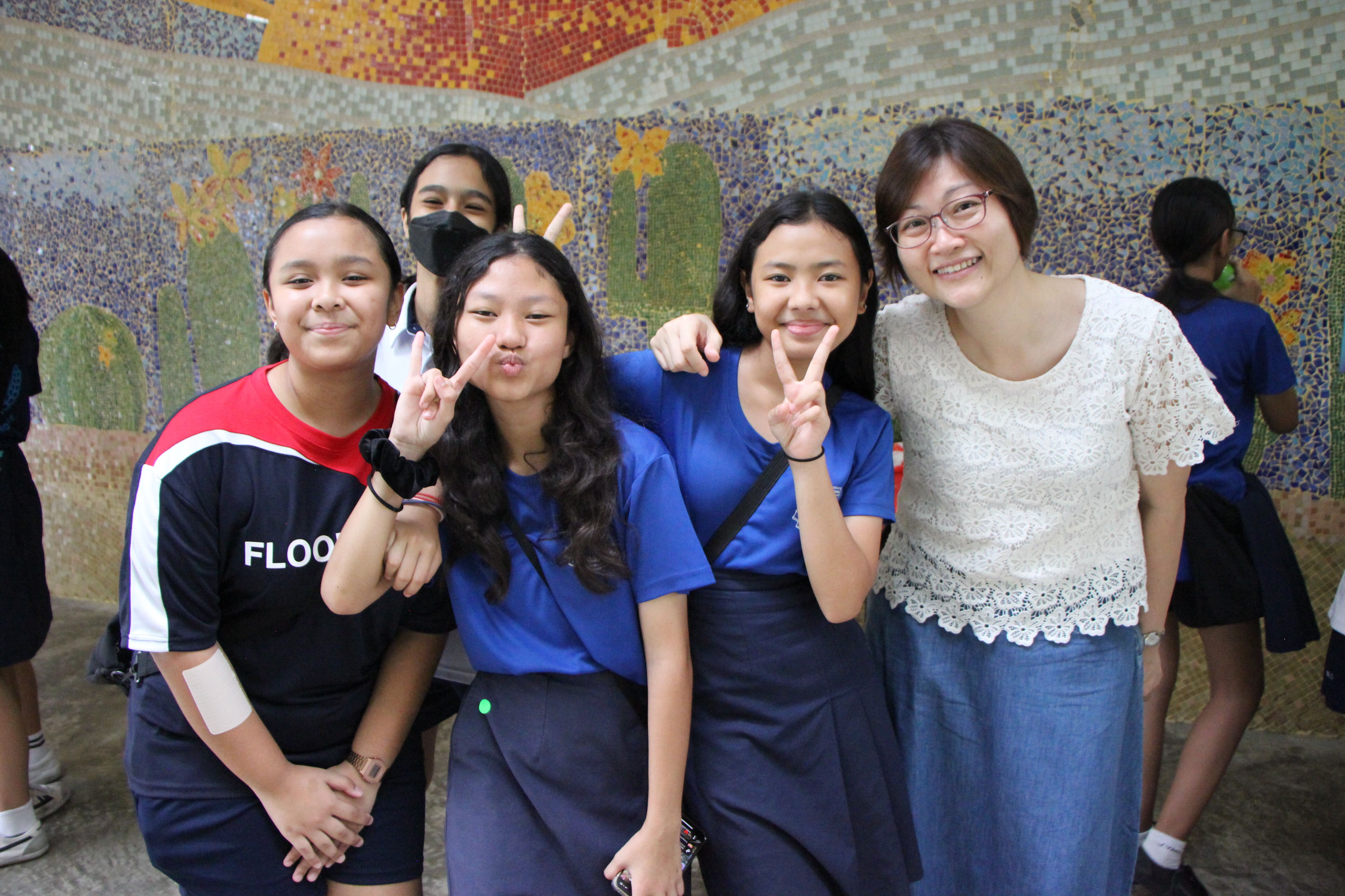 Five young women pose smiling in front of a colorful mosaic wall, two making peace signs.